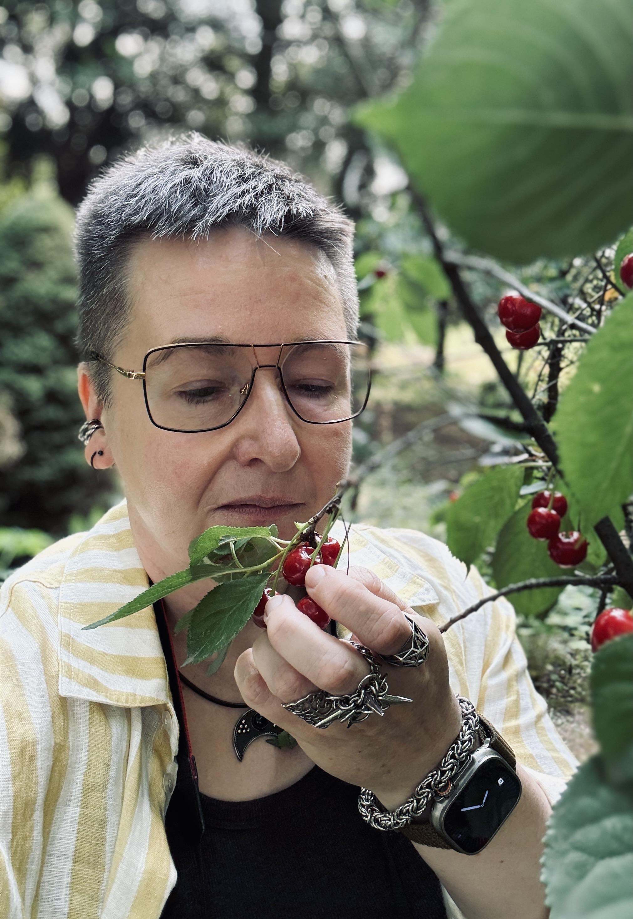 A person with short, gray hair is examining a cluster of red cherries while in a lush garden setting. They are wearing glasses and jewelry, and their focus is on the cherries held in their hand. The background is softly blurred with greenery.